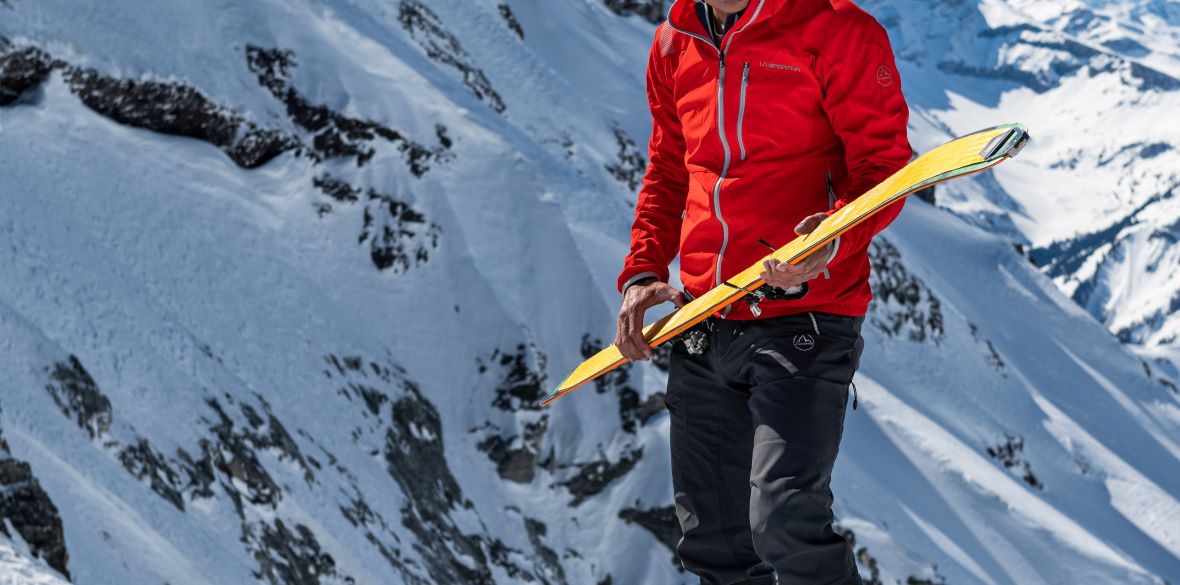 : Eine Person in roter Skijacke hält einen gelben Skifellstreifen in den Händen, während sie auf einem verschneiten Berghang steht.